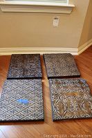 Four square metal plaques displayed on floor, showing different embossed patterns and metallic finish.