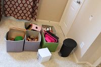 Four storage bins containing various toys such as Barbie dolls, toy cash register, furniture pieces, and a box and trash bin in corner of room.