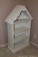 Photo of white wooden shelving unit shaped like a doll house with four shelves and chair rail trim.