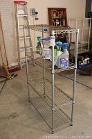 Full view of the wire utility shelving unit standing in a garage with various items on the shelves and background tools and ladder.