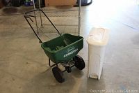 Overview of green Scotts Standard spreader with two wheels and metal handle alongside a white plastic trash bin with foot pedal opener.