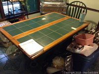 Dining table with green tile inset top and storage below. Two removable leaves on the table. Surrounded by furniture and household items.