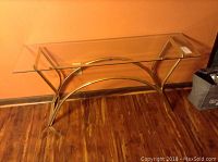 Front angled view of glass sofa table showing clear glass top with metal base against orange wall and wood floor.