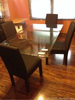 Four black upholstered dining chairs around a glass dining table in a room with wooden flooring and orange walls.