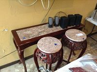 Sofa table and matching round end tables with marble tops and carved wood details in a room next to a yellow wall, with some audio speakers placed on top of the sofa table.