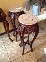 Two matching wooden plant stands with round beige marble tops and carved aprons on curved legs on tiled floor.