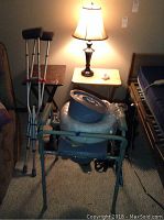 Full view of medical equipment and furniture in room including crutches, medical toilet chair, prosthetic leg, walking boot, dark base lamp, and wooden TV tables.