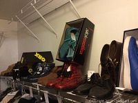 Photo showing multiple pairs of men's shoes and boots on a shelf, including shoes in original boxes labeled Geox and Siox Exotica, with colors such as red, teal, brown, and black.
