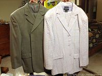 Two men's suit jackets hanging on hangers: light grey-green jacket and white pastel striped jacket