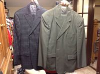 Two men's suit jackets on hangers, one in gray pinstripe and the other in solid gray-green, illustrating style and color variations.