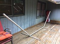 Wide angle side view of white metal hammock stand on wooden deck near siding and chair, showing full length and structure