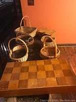 Chess board closed with visible wear, scratches on surface. Board dimensions approximately 31.25 x 15.75 x 5 inches. Seven wicker baskets in background of varying sizes and shapes with handles.