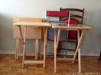 Four wooden folding TV tables with light wood finish and two folding chairs (one wooden with red vinyl seat and armrests, one metal with red vinyl seat and wooden backrest) leaning against wall on light wooden parquet floor.