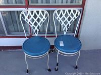 Two white metal chairs with lattice backrests and round blue seat cushions placed side-by-side outdoors.