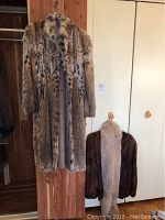 Two fur coats hanging beside a door, one long spotted pattern coat and one shorter brown coat with a beige collar hanging on it.