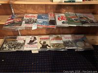 Photo showing a collection of 15 war-themed books and magazines lined up on wooden shelves. Includes titles on Berlin and the enemy, Welsh Civil War troops, the first frozen corps, and other war history topics.
