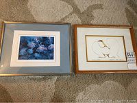 Photo shows two framed signed prints laid flat on a carpeted floor. Left print features multiple fish in blue tones. Right print features a dancing goose stylized artwork.