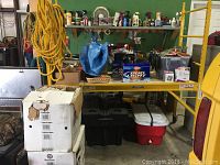Disassembled yellow metal scaffolding components, including the frame, plywood platform, and caster wheels shown upright against a workbench in a workshop area with other tools and items.