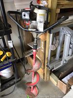 Full view of gas-powered earth auger powerhead with attached red auger bit leaning against shelving