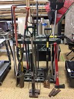 Front view of assorted yard tools inserted into black plastic storage stand, including rakes, shovels, digging forks and axes.