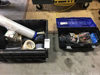Two black toolboxes containing power tools, tape rolls, plastic tubing, and accessories placed on a concrete floor.