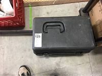 Closed black hard plastic carrying case on concrete floor, showing handle and metal latches.