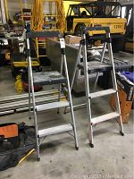 Front angle view of two aluminum step ladders standing side by side in a workshop, showing three steps and black top tray with paint marks.