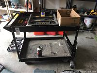Full shot of black metal rolling cart with lower and upper shelves, a canister on lower shelf, and tools on top shelf.