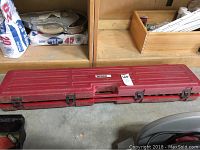 Closed view of the long red carrying case with latches, placed on a concrete floor among other workshop items.