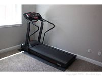 Full view of the treadmill showing the entire unit including the wide running belt, black frame, and handrails, placed against a wall in a carpeted room.