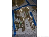 Wide view showing assorted silver plate and some stainless steel spoons, including miniature and larger serving spoons laid out on a cloth with blue lines.