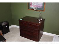 Wooden four drawer file cabinet with dark brown finish, two smaller drawers on top and two larger drawers below. Cabinet placed against green wall on carpeted floor with a silver adjustable desk lamp on top.
