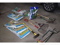 Wide view showing seven bags of potting soil and fertilizer alongside lawn and garden tools including rake, pitchfork, shovel, and axe all laid out on garage floor.