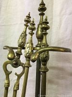 Close-up showing detail ornate brass handles of fire irons including poker, tongs, brush, and shovel, all hanging from brass stand.