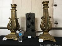 Photo showing two large gold-tone brass-style candle holders with ornate classical designs and one small electric fountain with three bell features in the center, water bottle for scale.