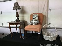Full view of upholstered peach chair with teddy bear on seat, adjacent dark wood side table with lamp on top, and white wicker basket to side.
