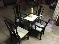 Dining set shown in garage setting with rectangular glass tabletop on black metal frame. Four wooden chairs with light patterned upholstery; two armchairs and two armless. Candle in metal holder placed at table center.