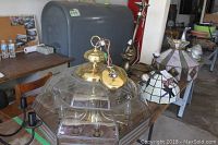 Wide view showing large clear segmented glass shade, brass-like chandelier with 6 arms, and multiple Tiffany style stained glass shades arranged on table.
