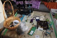 Overview of table with wicker basket, sunglasses, black velvet display stand with hanging necklaces, various jewelry items spread on table, and dark box with more jewelry inside.