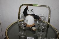 Close-up photo showing four clear glass liqueur glasses on the top glass shelf of brass-colored metal circular bar cart. Also visible is a stuffed panda toy beside the glasses, not part of the lot.