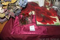 Artificial flower bouquet in tin vase, two red floral canvas art pieces and a small glass floral decor on a red cloth surface