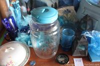 Overview photo showing large turquoise lidded glass jar, blue glass cups, dishes, feather boa, and purses in blue colors on wooden surface.