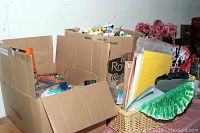 Boxes filled with craft supplies including spools of thread, beads, pipe cleaners, and felt pieces in a basket on a table along with artificial pink flowers in the background.
