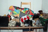 Wide shot of a table filled with various items including a colorful snow slider leaning against the wall, old phones, cables, black speakers, and boxes.