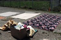 Photo shows several rugs laid on gravel ground including a large black rug with pink rose pattern and beige textured rug partially unrolled. Cardboard box and smaller cloth pieces visible.