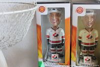 Photo showing three unopened Team Canada bobblehead dolls in their original packaging. One doll wears a white jersey, one a gold jersey, and one is in a black suit representing players and a coach.