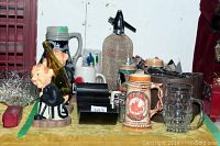 Photo showing novelty ceramic bottle holder, metal mesh seltzer bottle, German beer steins, assorted glassware and ice bucket.