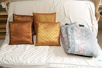 Four square pillows in bronze and gold tones with quilted diamond pattern covers and two folded blankets on white couch, showing minor wear.