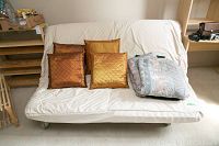 Futon placed upright with white mattress and four decorative pillows, mattress cover shows pilling and minimal stains, pillows orange and gold with quilt pattern.