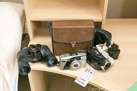 Lot displayed on wooden surface includes Bushnell binoculars, Vivitar camera, Olympus camera, camera case, power plugs, and straps.